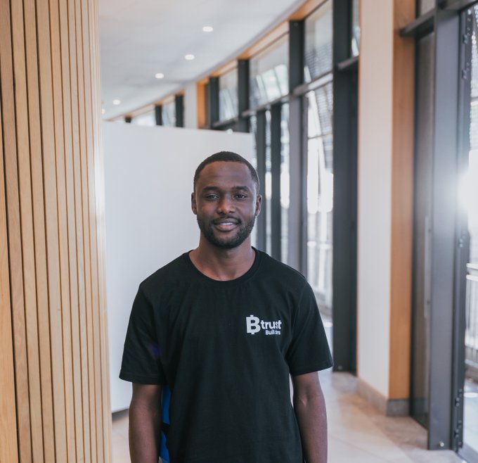 A smiling man with a short beard standing in a modern building with wood-paneled walls, wearing a black t-shirt with the 'Btrust Builders' logo.