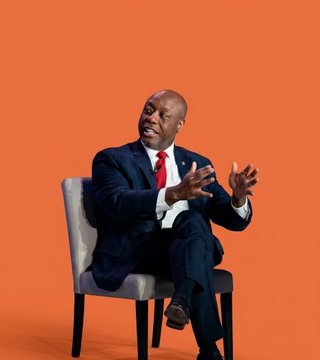 The image features a man dressed in a suit, sitting in an office chair with his hands outstretched as he speaks. The background of the image is orange, which contrasts with the man's dark suit and tie. There are no other objects or people visible in the image, making it a simple yet striking composition that highlights the man speaking.