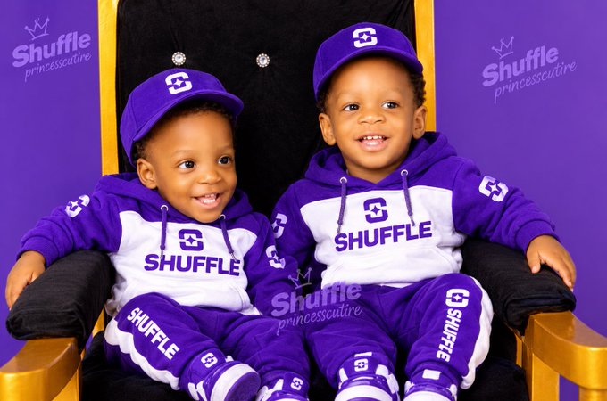 Two young children wearing matching purple and white 'Shuffle princesscutire' branded hoodies, sweatpants, and caps sit smiling in gold-framed chairs against a purple backdrop.