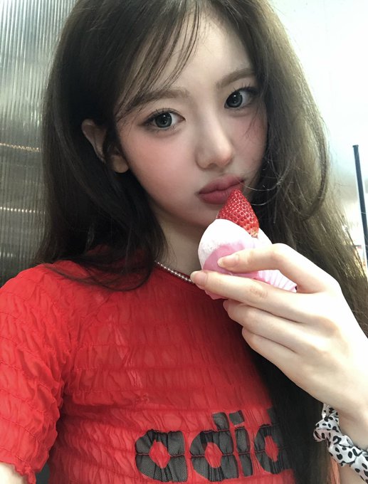 A girl with long dark hair takes a close-up selfie while holding a strawberry dessert near her mouth, wearing a red textured shirt with black lettering.