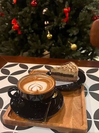 A cozy cafe scene featuring a black cup of cappuccino with latte art and a slice of chocolate cake on a wooden tray, with a decorated Christmas tree visible in the background.