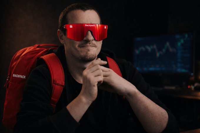 A man wearing red sunglasses labeled 'Backpack' and carrying a matching red 'Backpack' backpack, sitting in front of a computer monitor displaying financial charts.