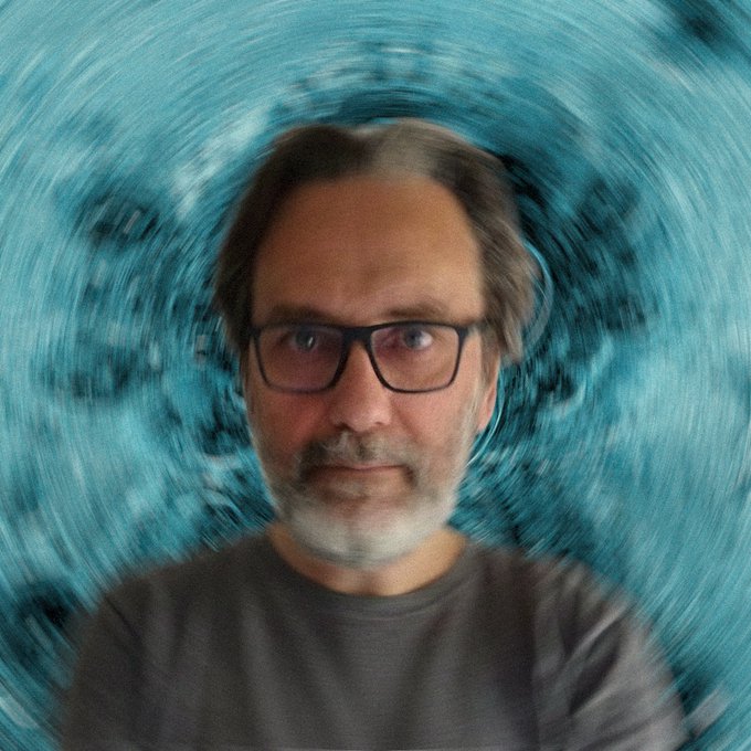 A portrait of a middle-aged man with glasses, a beard, and a gray shirt, with a swirling teal and blue abstract background behind him.