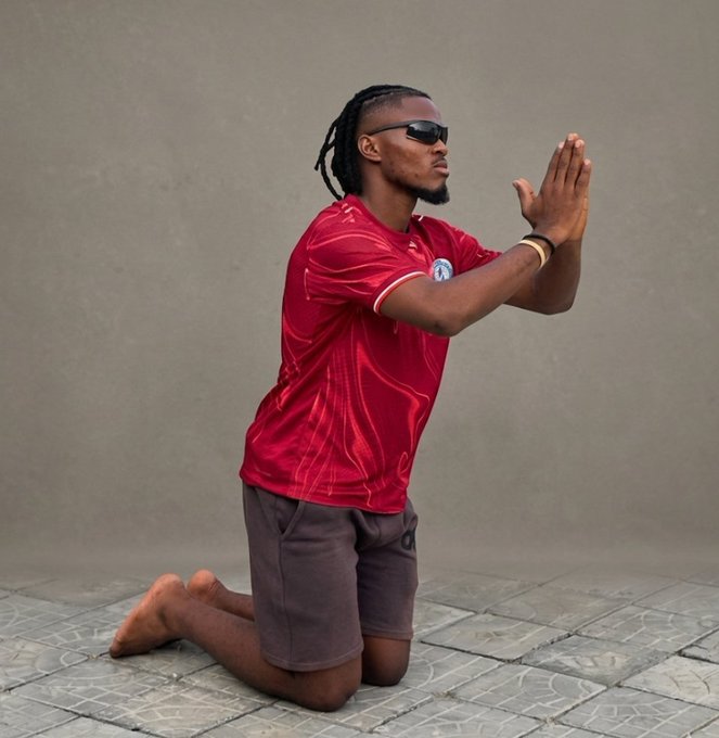 A man wearing sunglasses and a red jersey kneels on a tiled surface outdoors, pressing his palms together in a prayer position.