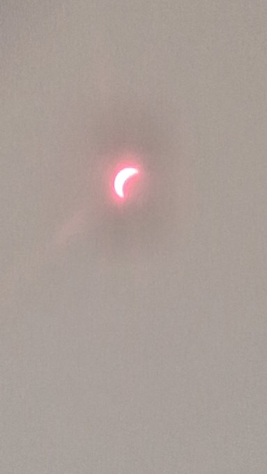 A photograph of a solar eclipse showing a red crescent sun against a hazy gray sky.