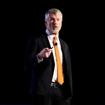 A man with gray hair and a goatee, wearing a dark suit and bright orange tie, stands against a black background, holding a small device while speaking. The image illustrates the accompanying message about Michael Saylor advocating for Bitcoin, emphasizing his confident stance as a presenter on stage.