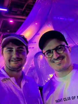 A close-up photo of two smiling young men posing together indoors, one wearing a cap and white shirt, the other wearing a cap, glasses, and a white t-shirt with 'YACHT CLUB OF N...' visible, lit by purple lighting.