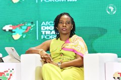 A woman wearing glasses and a yellow outfit speaks at the Africa Digital Finance Forum, with a green branded backdrop behind her.