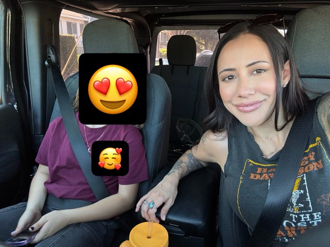 Two people sit in a car with a yellow smiley heart-eyes emoji covering one's face and a smaller blushing emoji with hearts below it, while the other smiles and holds an orange tumbler with a straw, wearing a black graphic tank top with partially visible text reading "THE" and "ON." The image captures a sweet mother-child moment, complementing the caption about how quickly children grow up, as they appear nearly the same height in the passenger seat.
