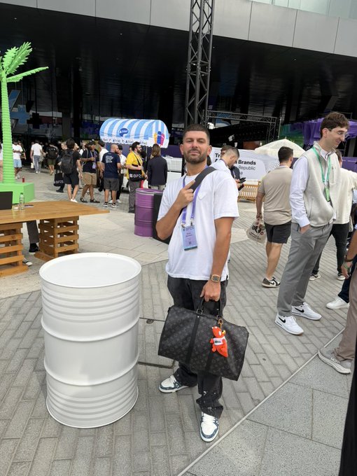The man in the image appears to be attending a crypto or Web3 conference, holding a Keepall-style travel bag (likely Louis Vuitton) with a small orange mascot keychain, while standing near a barrel and a "Brands" booth in the background. His outfit—a plain white t-shirt, jeans, and sneakers—contrasts with the high-end bag, mirroring the post's message about the crypto space being saturated with flashy projects ("vaporware") that lack substance or unity. The image suggests that even at industry events, appearances can be deceiving, reinforcing the call for genuine collaboration over hype-driven fragmentation.