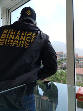 A man wearing a black cap and a black jacket with 'BINANCE' printed repeatedly in yellow on the back, standing by a window with a city view.