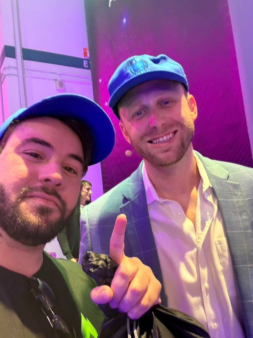 Two men wearing blue caps smiling and posing together in front of a stage with purple lighting, one pointing upward.