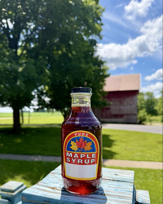 A glass bottle labeled 'PURE MAPLE SYRUP' with a Canadian maple leaf design sits on a wooden picnic table, with a red barn and green field in the background.