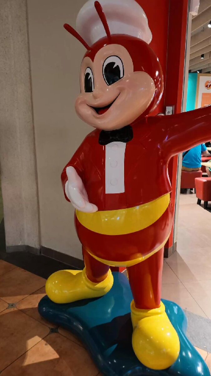 The image features a large Jollibee-themed statue of a cartoon character wearing red and yellow shoes. The statue is placed indoors near a wall, making it an eye-catching centerpiece for anyone passing by or visiting the establishment. The post suggests that one can use their stablecoins to purchase food from the restaurant, specifically ordering a meal at Jollibee with USDC or USDT.