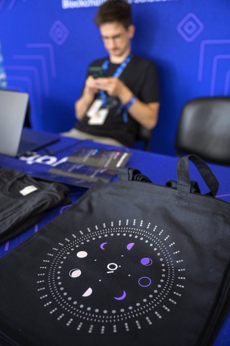 A black backpack featuring a moon phase calendar design sits on a table in the foreground, with a person in the blurred background using a phone. The setting appears to be a gaming or tech event with blue branded backdrops.