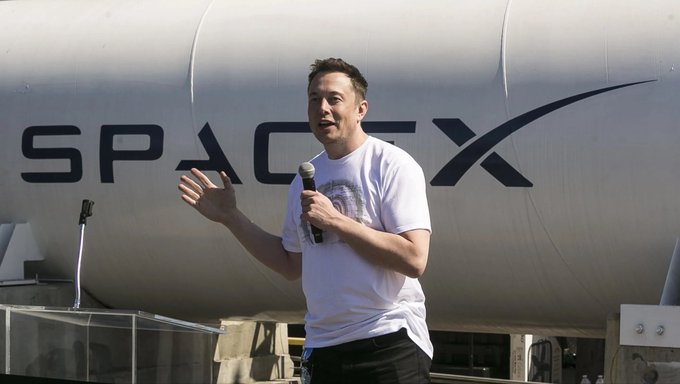 A man speaks into a microphone while gesturing with his hand, standing in front of a large structure displaying the "SpaceX" logo. The image relates to the post by visually associating the speaker with the SpaceX brand and the ticker $SPCX referenced in the headline about Elon Musk's AI predictions.