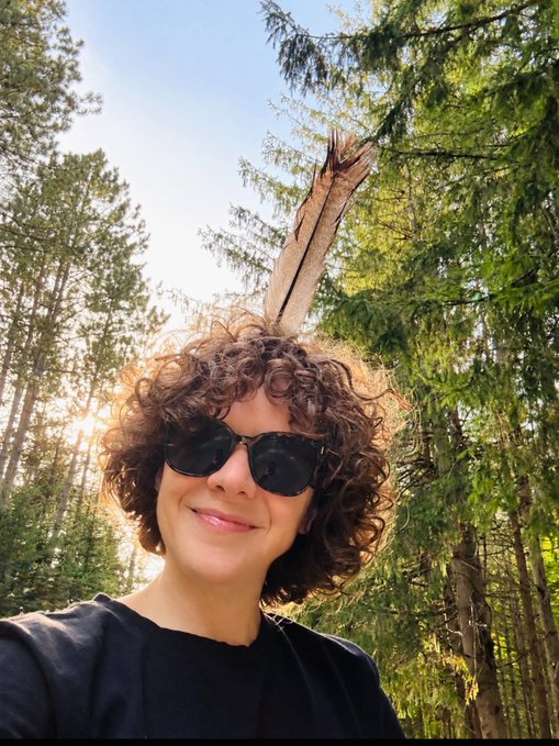 A selfie of a person with curly hair wearing sunglasses, standing in a sunlit forest with tall evergreen trees.
