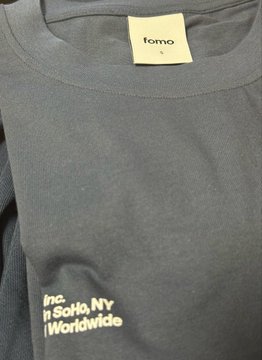 A gray "fomo" branded t-shirt featuring the brand's logo tag and "Designed in SoHo, NY | Distributed Worldwide" text, representing Fomo Labs Inc.'s crypto team merchandise.