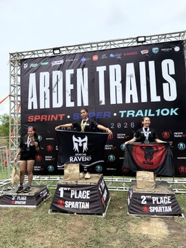 The image features three people standing on podiums, each holding a trophy or award. They are all wearing black shirts with "Arden Trails" written on them. The first person is located on the left side of the photo, while the other two individuals stand in the center and right side of the frame. All three of them have their arms raised high above their heads as they proudly display their achievements.