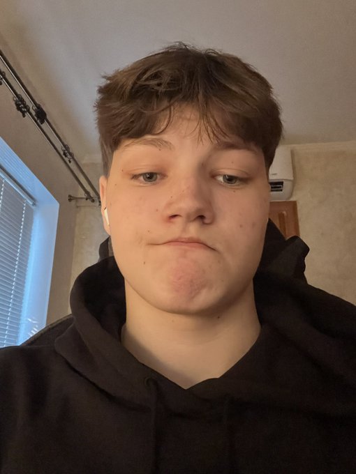 A selfie of a young person with short brown hair wearing a black hoodie and a white earbud, taken indoors with window blinds visible in the background.