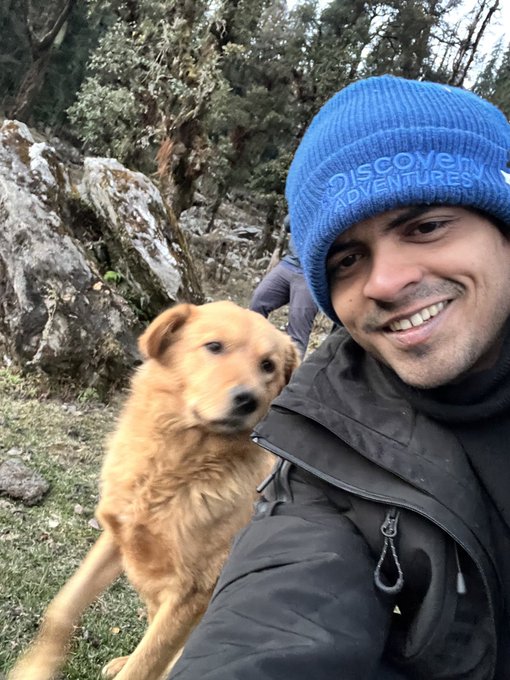 A smiling man in a blue beanie with the words 'DISCOVER ADVENTURES' takes a selfie alongside a golden-furred dog in a wooded, rocky area.