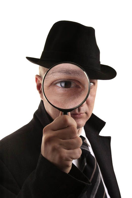 A man wearing a black fedora, black coat, and striped tie holds a magnifying glass up to his right eye against a stark white background.