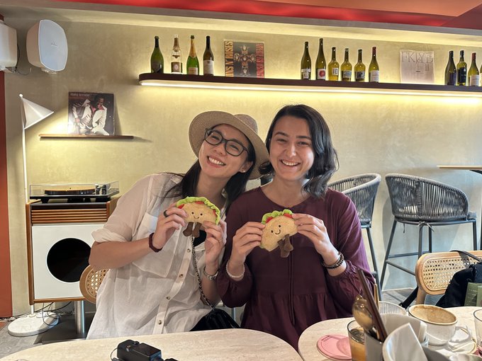 Two smiling women sitting at a restaurant table, each holding up a plush taco toy, with bottles displayed on a shelf in the background.