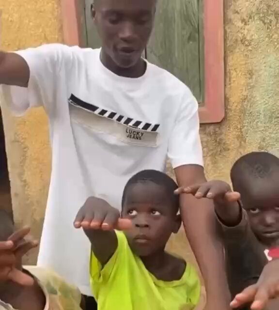 The image shows a man wearing a white t-shirt with black text, standing next to two children who are holding his arms up. The background of the photo features a yellow wall that has "Nigerians will do anything for a ." written on it in black letters. This suggests that the scene might be related to an event or situation where Nigerians have come together and demonstrated their willingness to help others, regardless of the circumstances.