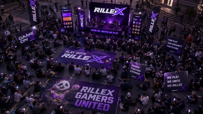 An aerial view of a large nighttime gathering for the gaming platform 'RILLEX', featuring banners with slogans like 'GAME ON. UNITED WE PLAY', 'PLAY WITH RILLEX', and 'RILLEX GAMERS UNITED', with a crowd seated on the ground facing a stage.