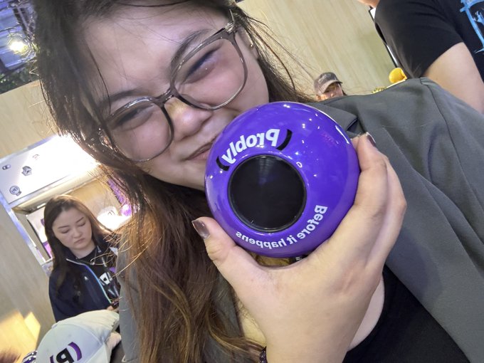 A young woman with glasses smiles while holding up a purple Prodigy ball labeled 'Beauty responds' near her face.