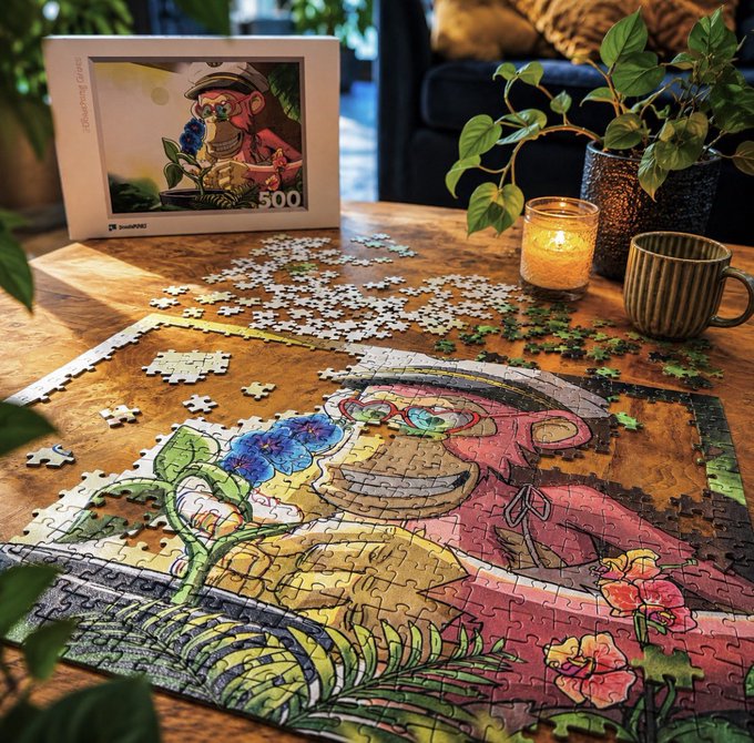 A partially completed 500-piece jigsaw puzzle by 'magicdoughart' showing a colorful illustration of a person with flowers, sitting on a wooden table with scattered pieces, a candle, a green mug, and the puzzle box in the background.