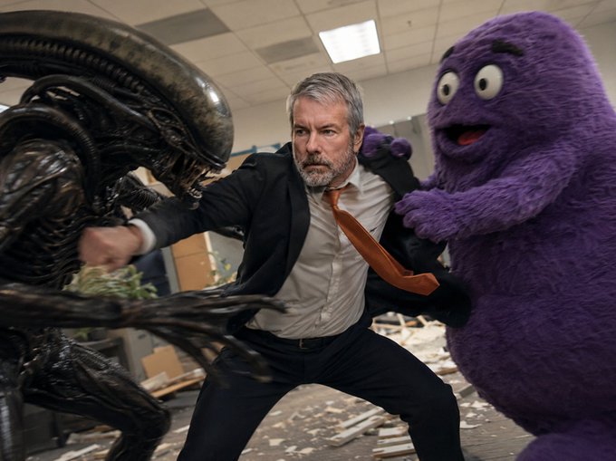 A man in a suit and orange tie stands defensively between a Xenomorph from Alien and the purple Grimace character in an office setting.