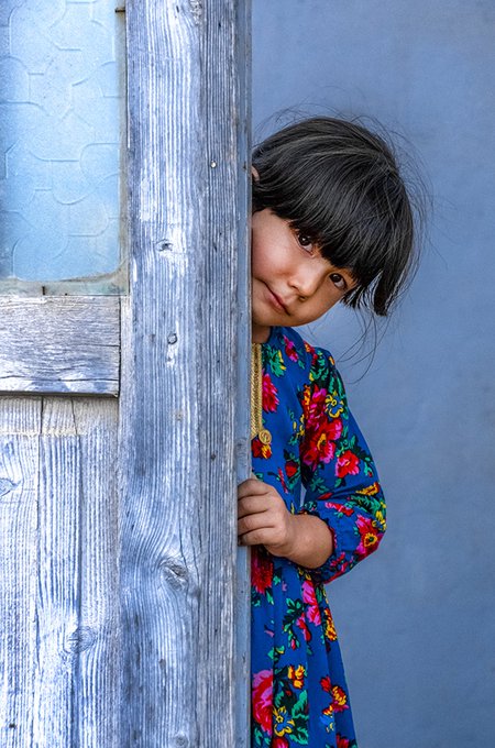 A young child with dark hair peeks out from behind a weathered wooden door against a blue wall, wearing a vibrant blue floral dress.