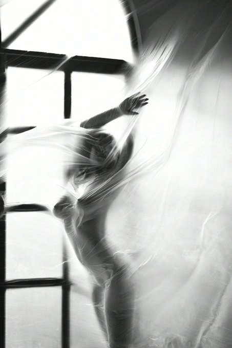 A lone figure is silhouetted behind a sheer, translucent curtain, their form partially obscured by the rippling fabric. The image is captured in black and white, with strong contrasts between light and shadow.