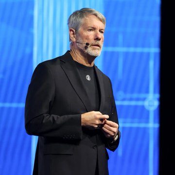 In the image, a man wearing a suit is standing on stage with his hand over his mouth while giving a speech or presentation, possibly addressing Bitcoin mining. The background features a blue wall with white text that provides an explanation about the current status of bitcoin mining. The speaker's facial expression suggests he may be explaining something serious or important.