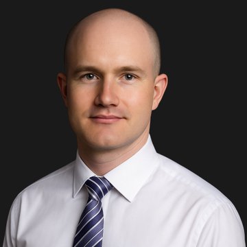 A headshot of a bald man in a white dress shirt and navy striped tie, presumably Coinbase CEO Brian Armstrong, shown against a dark background—illustrating the figure whose X profile picture change inadvertently triggered a 37x surge and 85% crash in the $BRIAN meme coin on Base.