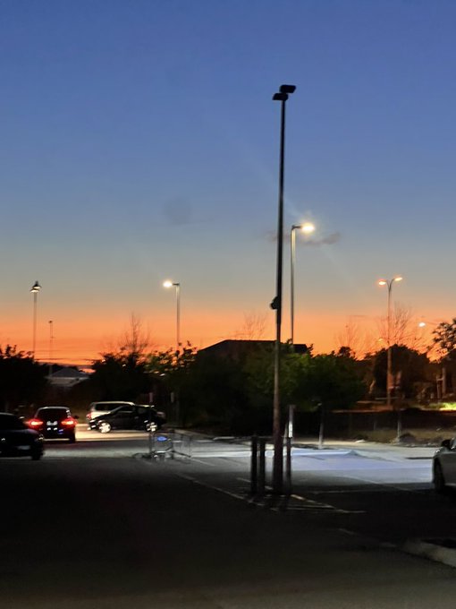 A parking lot at dusk with illuminated street lamps, parked cars, and a vibrant orange sunset fading into a deep blue sky.