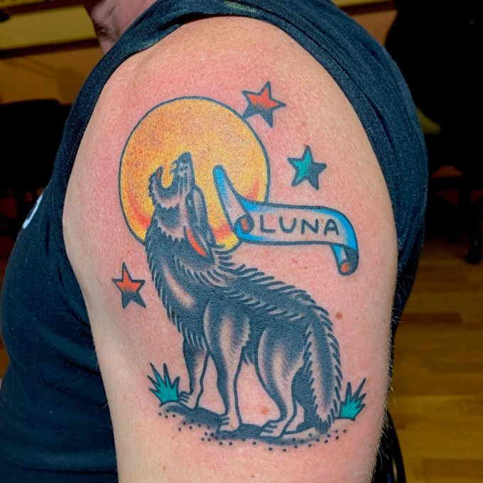 A close-up photograph of a tattoo on a person's upper arm depicts a howling wolf silhouetted against a bright orange moon, with a banner reading 'LUNA' and scattered stars.