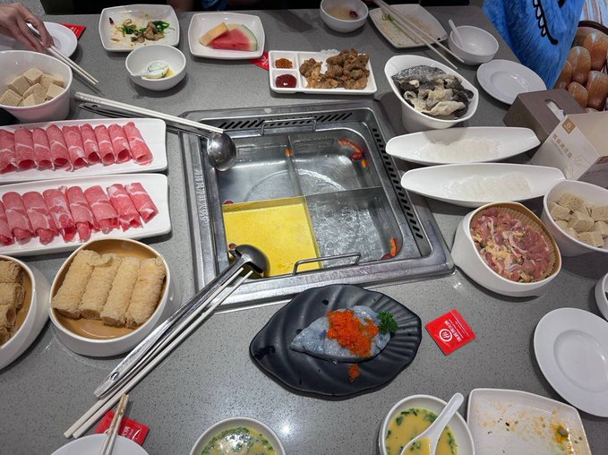 An overhead view of a table set for a hot pot meal, featuring plates of rolled meat, dumplings, vegetables, dipping sauces, and a divided pot with broth on a built-in stove.