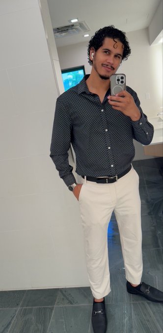 A man with curly hair takes a mirror selfie wearing a dark patterned button-up shirt, white pants, a black belt, and black loafers.
