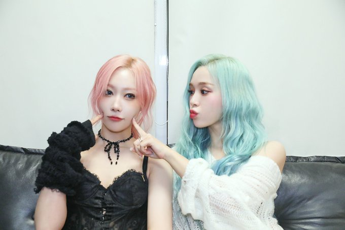 Two women with pink and mint-green hair pose together on a black couch, with one playfully poking the other's cheeks.
