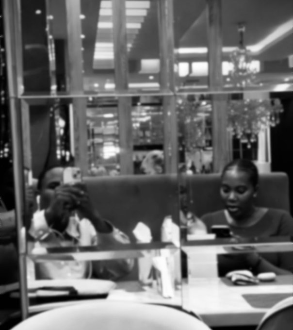 Black and white photo of two people at a restaurant, one taking a mirror selfie with an iPhone.