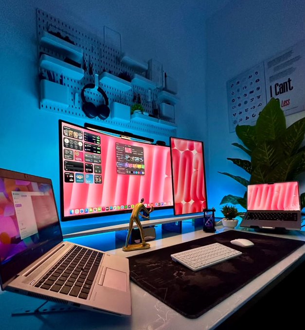 A modern desk setup featuring dual monitors and a laptop with pink/red abstract wallpapers, illuminated by blue and pink LED lighting, with a small figurine and plants on the desk.