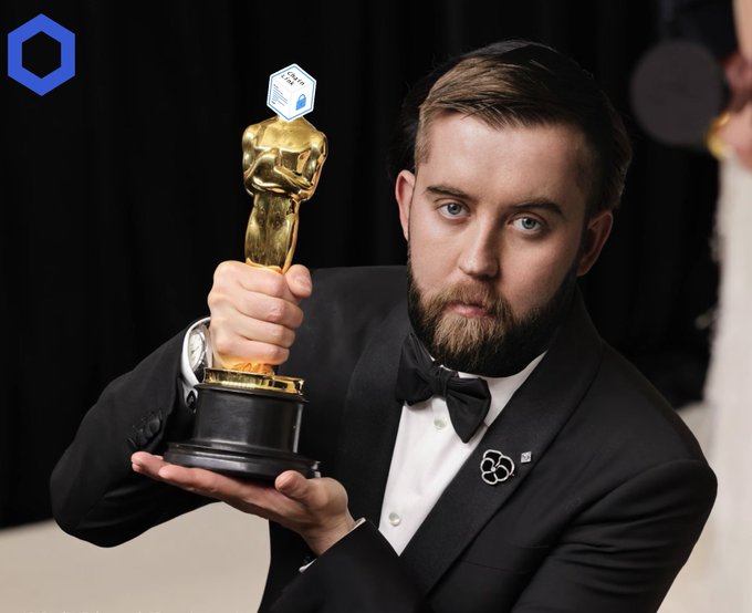 A bearded man in a black tuxedo with a bow tie holding a golden trophy at what appears to be an awards ceremony, with a blue hexagonal logo in the corner.