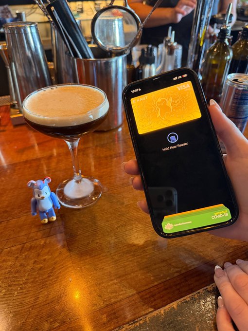 Photo of a hand holding a smartphone displaying an eshafl.io digital vaccination/health passport for COVID-19, with the time 18:07, positioned next to an espresso martini cocktail and a small blue figurine on a wooden bar.