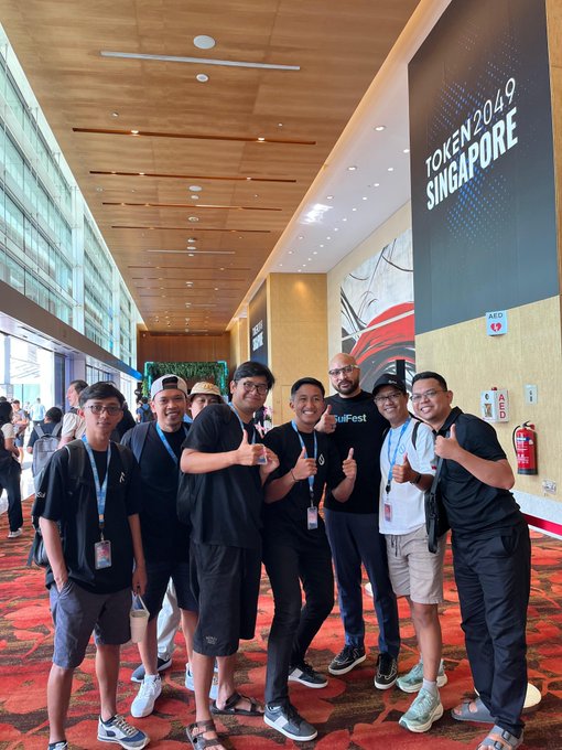 A group of smiling people pose together at an event, standing in front of a large banner that reads 'TOKEN2049 SINGAPORE'.