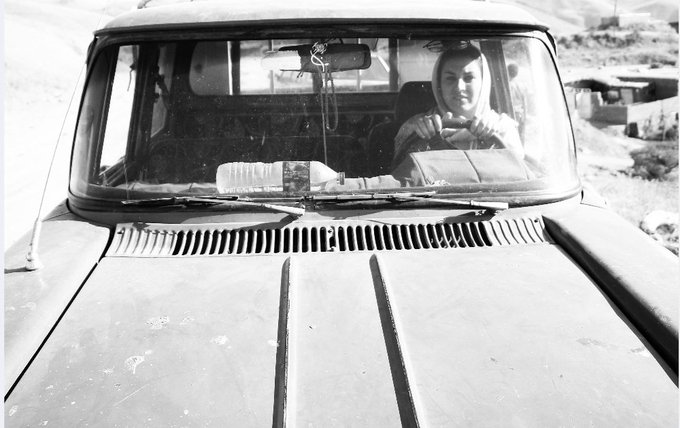 A black and white photograph of a woman sitting in the driver's seat of an old truck, looking through the windshield with her hands on the steering wheel.