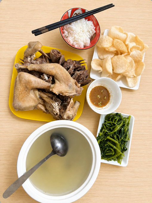 A meal laid out on a wooden table features steamed chicken pieces, white rice with chopsticks, prawn crackers, a dipping sauce, sautéed greens, and a bowl of clear soup with a spoon.