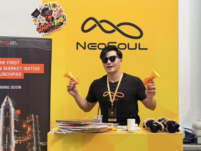 A man wearing sunglasses and a black t-shirt stands behind a vibrant yellow "NeoSoul" branded booth, holding up two orange toy airhorns while surrounded by merchandise including stickers and plushies, with a poster promoting "THE FIRST MARKET-NATIVE LAUNCHPAD" visible to the left.