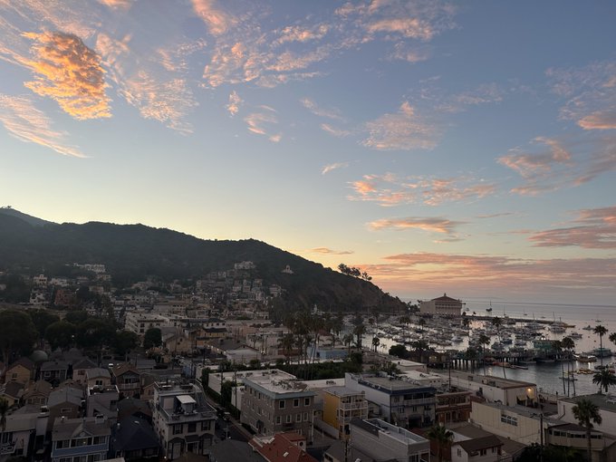 A scenic coastal town at sunset shows a harbor filled with boats, houses built into the hillside, and pink-tinged clouds in the sky.